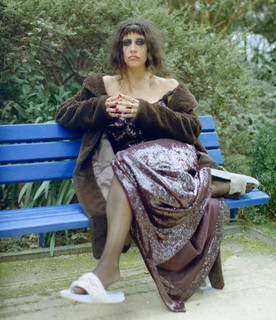 L'artiste Gildaa assise sur un banc dans la nature, yeux maquill&eacute;s de noir, m&eacute;lange de robe de soir&eacute;e et de v&ecirc;tements d'int&eacute;rieur (claquettes, manteau...) et cheveux d&eacute;braill&eacute;s