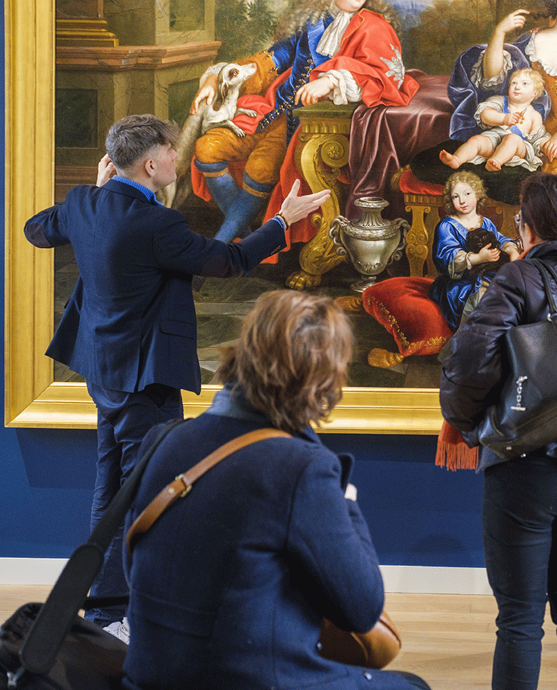 Un m&eacute;diateur montrant le tableau de Pierre Mignard, o&ugrave; l'on peut voir en arri&egrave;re-plan le monarque assis et deux de ses enfants dans un d&eacute;cor tr&egrave;s opulent